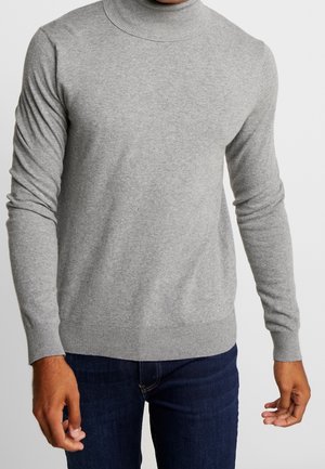 Homme portant un pull à col roulé gris clair et un jean bleu foncé, debout avec les bras détendus le long du corps.
