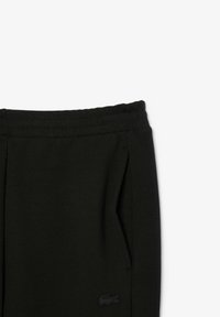Un pantalon de jogging noir avec une taille élastique, des poches latérales et un petit logo noir sur la cuisse gauche. Texture lisse, design décontracté.