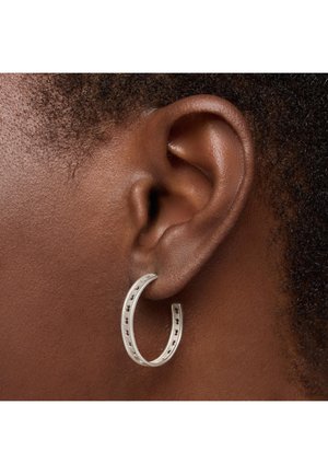 Pendiente de aro plateado con recortes, diseño circular. Está sujeto a un tono de piel oscura, mostrando el lóbulo de la oreja y el cabello texturizado en el fondo.