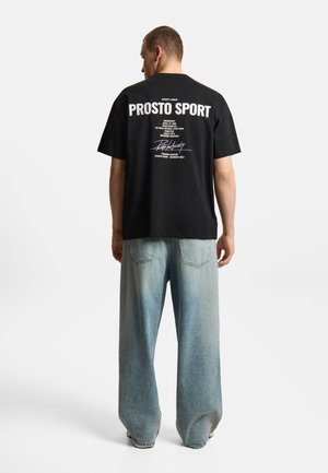 Mężczyzna w czarnej koszulce "PROSTO SPORT" i luźnych jasnoniebieskich dżinsach, stoi tyłem na jednolitym białym tle.
