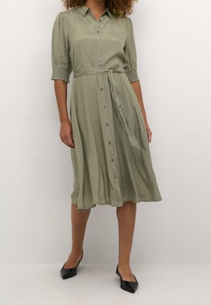 Femme portant une robe boutonnée vert olive longueur genoux avec ceinture à nouer et manches smockées mi-longues, associée à des ballerines noires pointues.