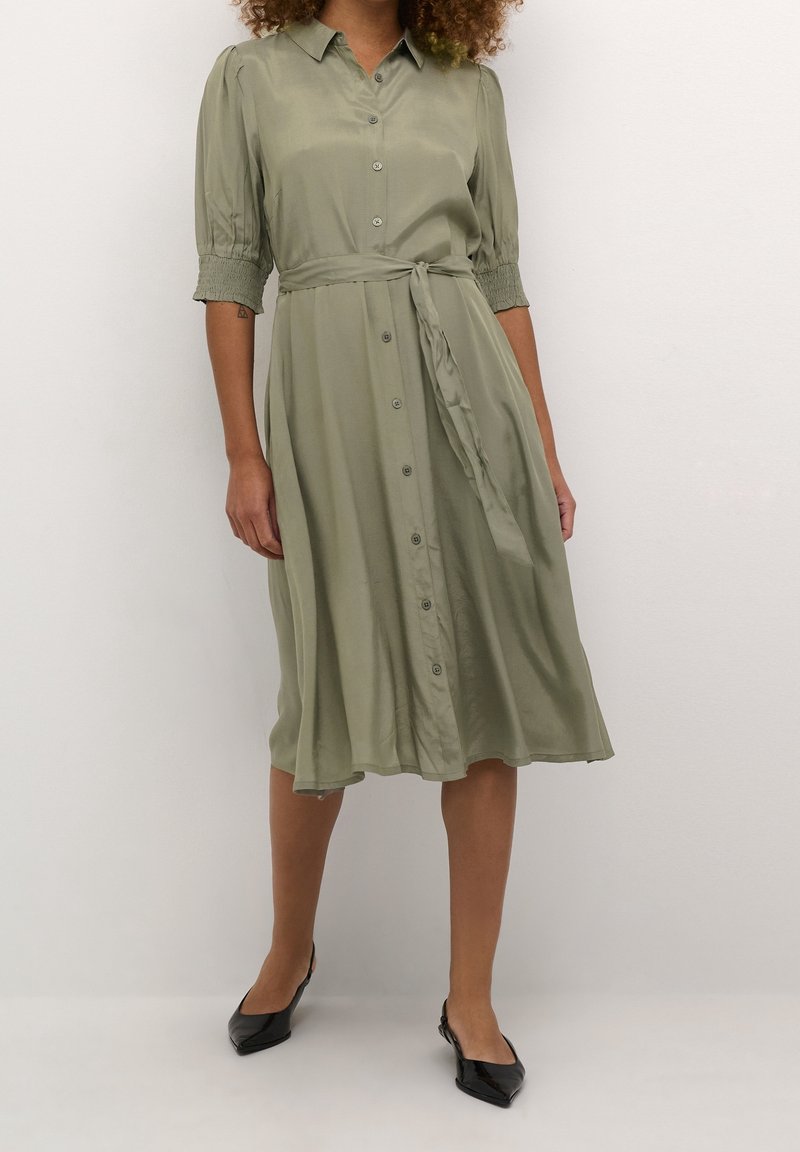 Femme portant une robe boutonnée vert olive longueur genoux avec ceinture à nouer et manches smockées mi-longues, associée à des ballerines noires pointues.
