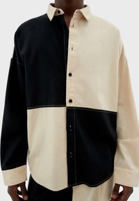 Camisa de manga larga con un diseño dividido en negro y beige, con costuras visibles, botones en la parte delantera y un corte relajado.