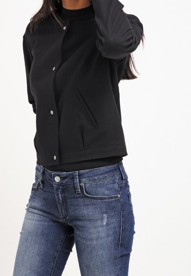 Femme portant une veste noire boutonnée sur un haut noir et un jean bleu, le bras droit levé près de la tête.