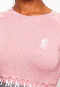 Růžový sportovní top s přiléhavým střihem, měkkou texturou, kulatým výstřihem a bílým logem na prsou a vzorovaným pasem dole.