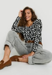 Haut noir à manches longues avec un motif courbé blanc, associé à un jean gris taille haute et des mocassins en suède marron, avec une ceinture marron.