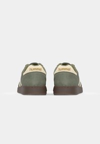 Grüne Wildledersneaker mit beigen Akzenten, ausgestattet mit einer braunen Gummisohle und drei dunklen Streifen. Auf der Rückseite befindet sich das goldene "hummel"-Logo.