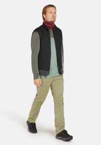 Schwarze gesteppte Weste über einem grünen Langarmhemd, kombiniert mit hellgrünen Hosen und schwarzen Wanderschuhen. Enthält einen weinroten Halswärmer.