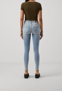 Femme portant un jean skinny taille haute bleu clair avec des ourlets effilochés, un top ajusté manches courtes vert olive et des sandales noires à talons épais.