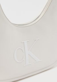 Λευκή δερμάτινη τσάντα με ασημένιο φερμουάρ και ανάγλυφο λευκό λογότυπο "cK" στο μπροστινό μέρος.