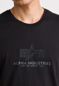 Svart bomulls t-skjorte med en hevet, teksturert logo i mørkegrått med "ALPHA INDUSTRIES" og tekst om sted trykket under. Rund hals.