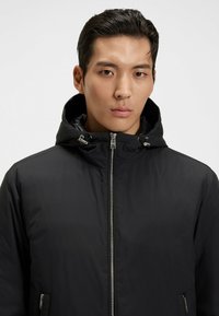 Ung man med kort svart hår iförd en svart huva-jacka som är dragkedjad, vänd framåt mot en enfärgad vit bakgrund.