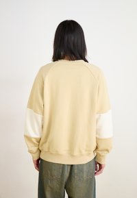 Felpa beige con maniche lunghe a raglan, caratterizzata da una decorazione bianca su una manica. Tessuto morbido, vestibilità comoda, orlo e polsini a coste ben visibili.