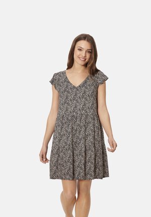 Joven sonriente con un vestido de manga corta con patrón en blanco y negro y escote en V, de pie contra un fondo blanco liso.