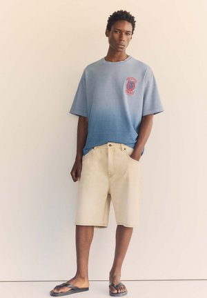 Jeune homme debout contre un mur beige portant un t-shirt dégradé bleu, un short beige et des tongs noires avec une main dans la poche.