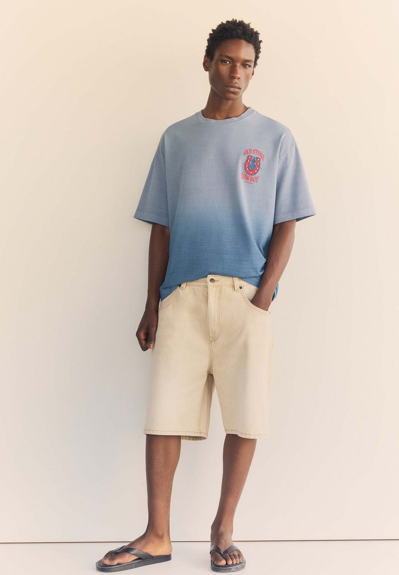 Jeune homme debout contre un mur beige portant un t-shirt dégradé bleu, un short beige et des tongs noires avec une main dans la poche.