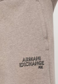 Tuvumā redzamie gaiši brūni auduma bikses ar sānu kabatu un melnu izšūtu "ARMANI EXCHANGE N.Y." logo netālu no kabatas.