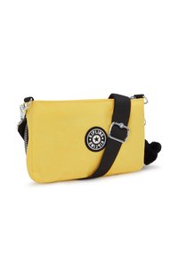 Keltainen nylonselkäpussi, jossa on musta hihna, ja jossa on pyöreä logo-ommel sekä musta pom-pom -yksityiskohta. Vetoketjusulku toisella puolella.