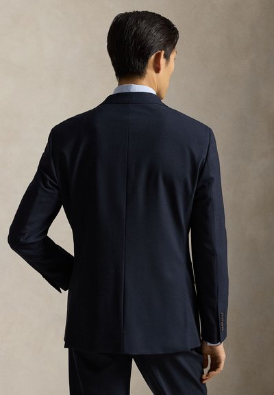 Μπλε σκούρο σακάκι κοστουμιού, με ραμμένο σχέδιο και δομημένους ώμους, γιακάδες με σχισμή και μία μονή σχισμή στο πίσω μέρος. Λείο υφή, με τέσσερα κουμπιά στα μανίκια.