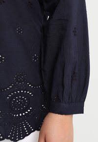 Chemise à manches longues bleu marine ornée de motifs floraux brodés et de découpes au niveau de l'ourlet et des manches. Tissu doux et texturé.