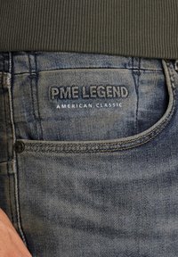 Jeans en denim avec une teinte bleue délavée, arborant "PME LEGEND" brodé sur la poche arrière, rehaussé de surpiqûres orange et d'un rivet.