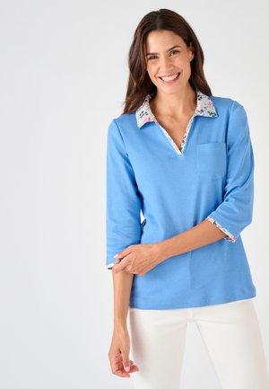 Lächelnde Frau mit dunklen Haaren, die ein hellblaues Oberteil mit floralem Kragen und Manschetten trägt, richtet ihren Ärmel, steht vor weißem Hintergrund.