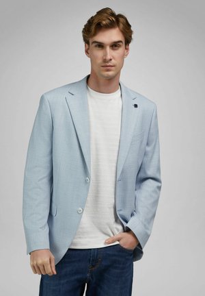 Junger Mann mit hellbraunem Haar trägt ein hellblaues Sakko über einem weißen, strukturierten Hemd und blauen Jeans, steht mit einer Hand in der Tasche.