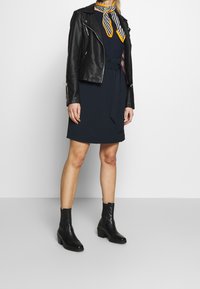 Mujer vestida con una chaqueta de cuero negra sobre un vestido azul marino con cinturón, botas negras hasta el tobillo y una bufanda de cuello a rayas amarilla, blanca y negra.