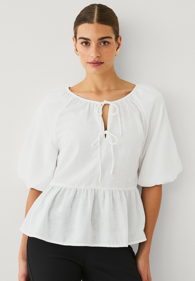 Bílá peplum blůza z lehkého materiálu. Má kulatý výstřih s mašlí, rourové rukávy a volánkový lem.
