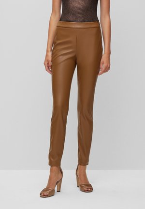 Schmal geschnittene braune Lederhose, getragen mit beigefarbenen Sandalen mit hohem Absatz, kombiniert mit einem ärmellosen braunen metallicfarbenen Top vor einem schlichten Hintergrund.