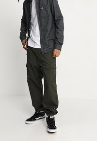 Camisa gris con estampado y botones sobre una camiseta blanca, combinada con pantalones cargo verde oliva y zapatillas negras con detalles blancos.