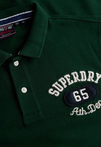 Polo vert en tissu texturé avec un col boutonné. Logo brodé "SUPERDRY 65" avec un patch d'accent bleu marine.