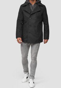 Cappotto peacoat nero con grandi risvolti e bottoni doppiopetto, sopra felpa grigia testurizzata; abbinato a jeans skinny grigi e sneakers bianche.