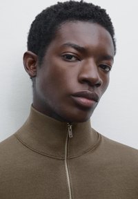 Young Black man with short curly hair wearing a brown zip-up sweater, looking slightly to the side against a plain light background.