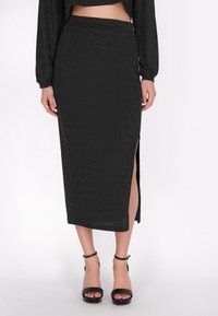 Czarna midi spódnica z bocznym rozcięciem, teksturowana tkanina z metalicznymi drobinkami i dopasowanym pasem w talii. W zestawie z czarnymi sandałami na obcasie.