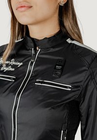 Giacca da corsa nera realizzata in nylon morbido, con dettagli bianchi, colletto alto, due tasche con zip e dettagli di branding ricamati.