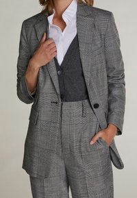 Szary marynarka w kratę z klapami, dwoma przednimi kieszeniami i pojedynczym zapięciem na guzik, nałożona na szary kamizelkę i białą koszulę.