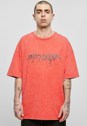 Rød oversized t-skjorte laget av bomull med en lett falmet tekstur. Har svart grafisk tekst over brystet. Korte ermer og ribbestrikket krage.