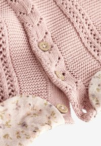 Gros plan d'un tissu tricoté rose avec motif tressé, boutons en bois et poignets doublés de tissu floral.