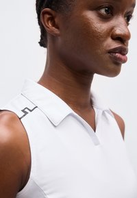 Κοντινό πλάνο ενός ατόμου που φοράει λευκό πουκάμισο χωρίς μανίκια με γιακά και μαύρη γραφική λεπτομέρεια στον ώμο.