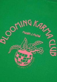Žalia marškinėliai su rožinės spalvos tekstu ir piešiniu, kuriame pavaizduota vazoninė gėlė. Tekstas skelbia "ŽIEDINTI KARMOS KLUBAS" ir "GĖLĖ & SROVĖ."