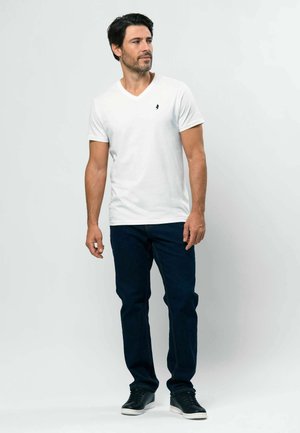 Camiseta blanca de algodón de manga corta con cuello en V y un logo azul marino. Combinada con vaqueros de mezclilla azul oscuro y zapatillas negras, posando frente a un fondo claro.