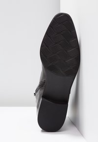 Semelle de botte noire avec motif ondulé et talon texturé, botte appuyée contre un mur d'angle blanc sur une surface lisse.