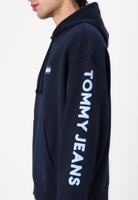 Persona vistiendo una sudadera con capucha azul marino con "TOMMY JEANS" impreso verticalmente en blanco en la manga y un pequeño logo en el pecho.