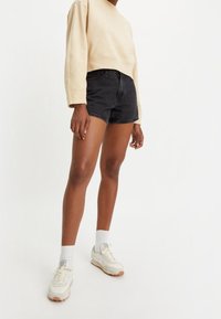 Sudadera beige recortada combinada con pantalones cortos de mezclilla negros de tiro alto. Zapatillas blancas con detalles en gris y calcetines gris claro.