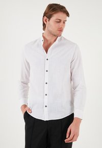 Camisa blanca de lino de mangas largas con cuello alzado, textura sutil y botones oscuros en la parte delantera. Combinada con pantalones negros.