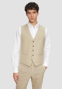 Beige Weste aus einem strukturierten Stoff, mit einem V-Ausschnitt, fünf braunen Knöpfen und zwei Taschen, kombiniert mit einem weißen Hemd.