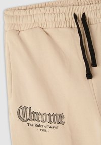 Jasno beżowe spodnie dresowe z elastyczną talią i czarnymi sznurkami. Na lewej udzie znajduje się wyraźny czarny logo i tekst.