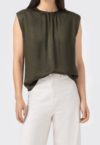 Mörk olivgrön ärmlös topp med rund halsringning och rynkade detaljer framtill; bärs med ljusa byxor.