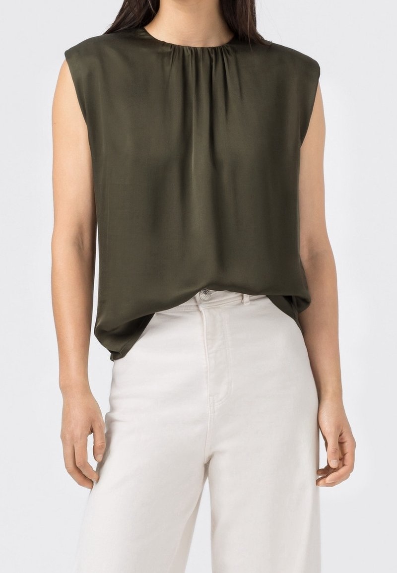 Mörk olivgrön ärmlös topp med rund halsringning och rynkade detaljer framtill; bärs med ljusa byxor.